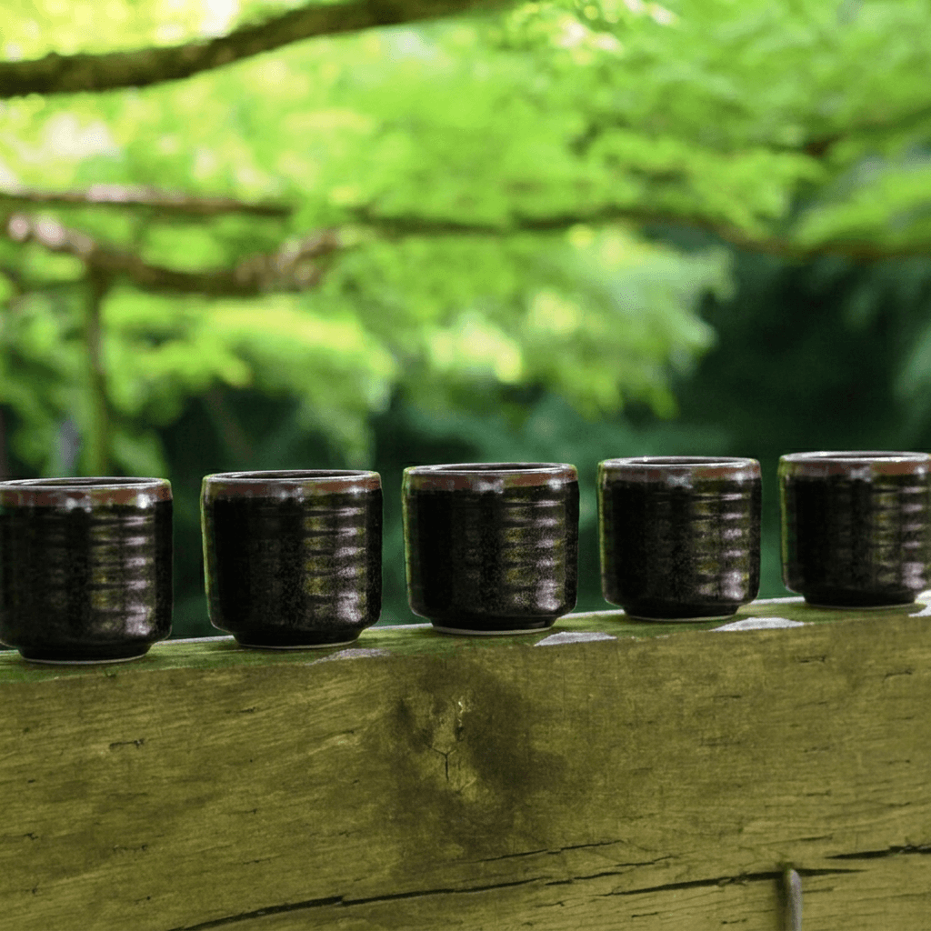Five small dark glazed ceramic cups lined up on a weathered wooden railing with a blurred green forest background.