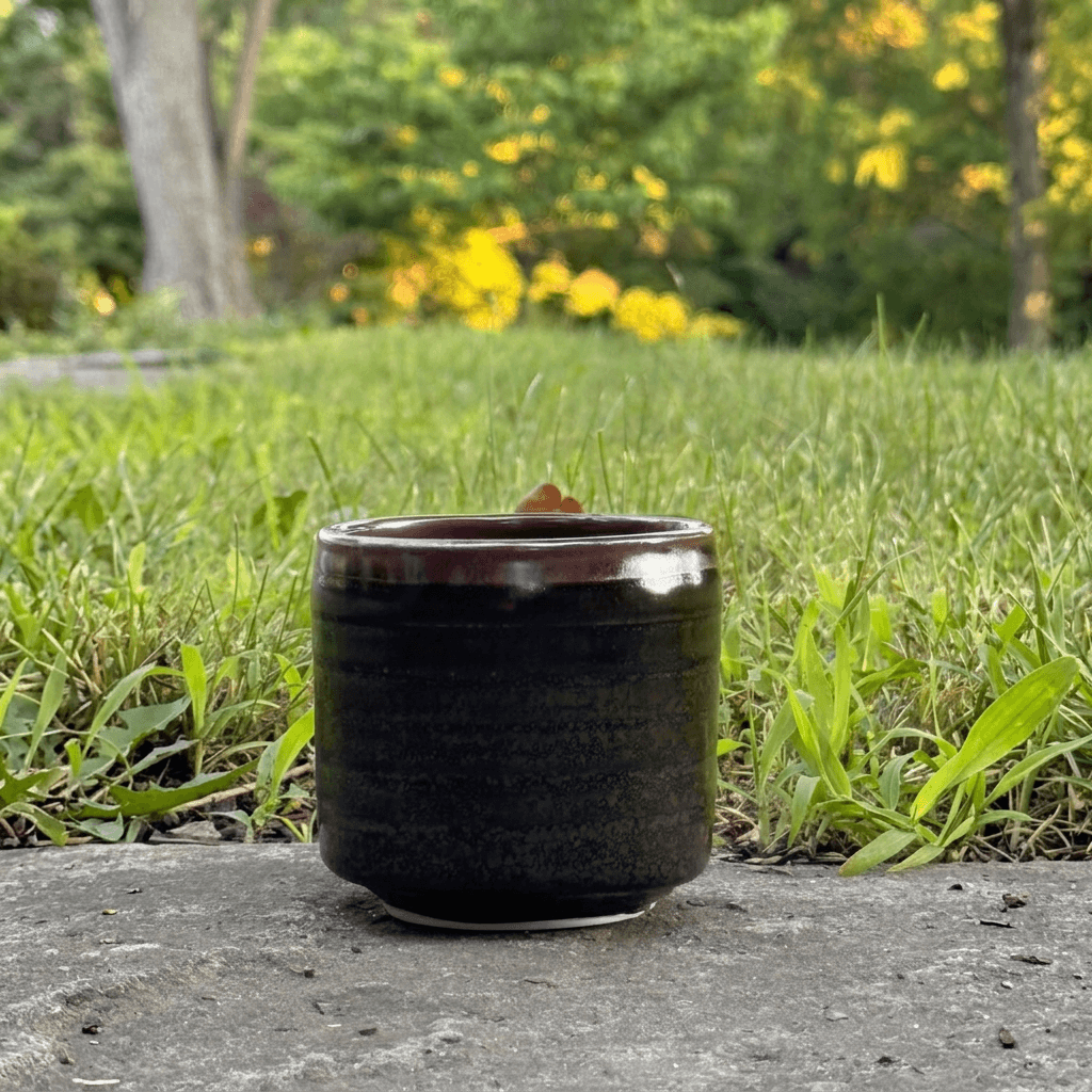 A small dark glazed ceramic cup or candle holder sits on a stone surface outdoors, with green grass and blurred trees in the background.