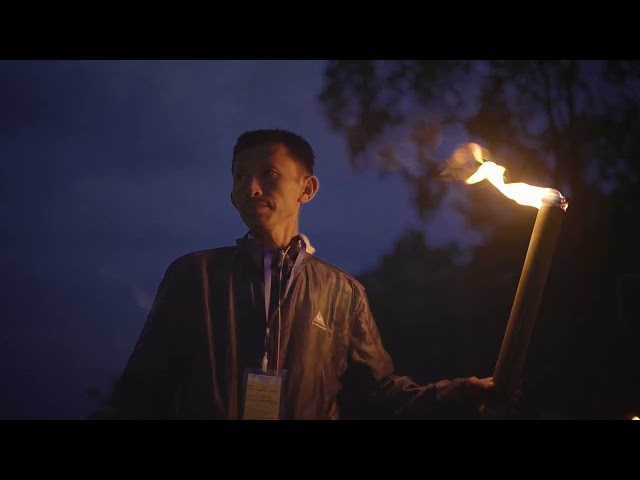 A man at night holding a lit torch, wearing a dark jacket and a lanyard with an ID badge, with a dark outdoor background.