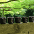 Five small dark glazed ceramic cups lined up on a weathered wooden railing with a blurred green forest background.