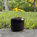 A small dark glazed ceramic cup or candle holder sits on a stone surface outdoors, with green grass and blurred trees in the background.