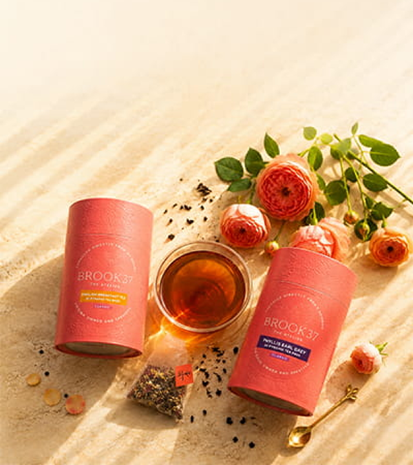 Two pink cylindrical tubes of BROOK37 tea, one labeled Rose Gold and one labeled Vanilla Bean + Rose, arranged on a sandy surface with a glass of amber tea, loose tea bags, rose petals, and green leaves.