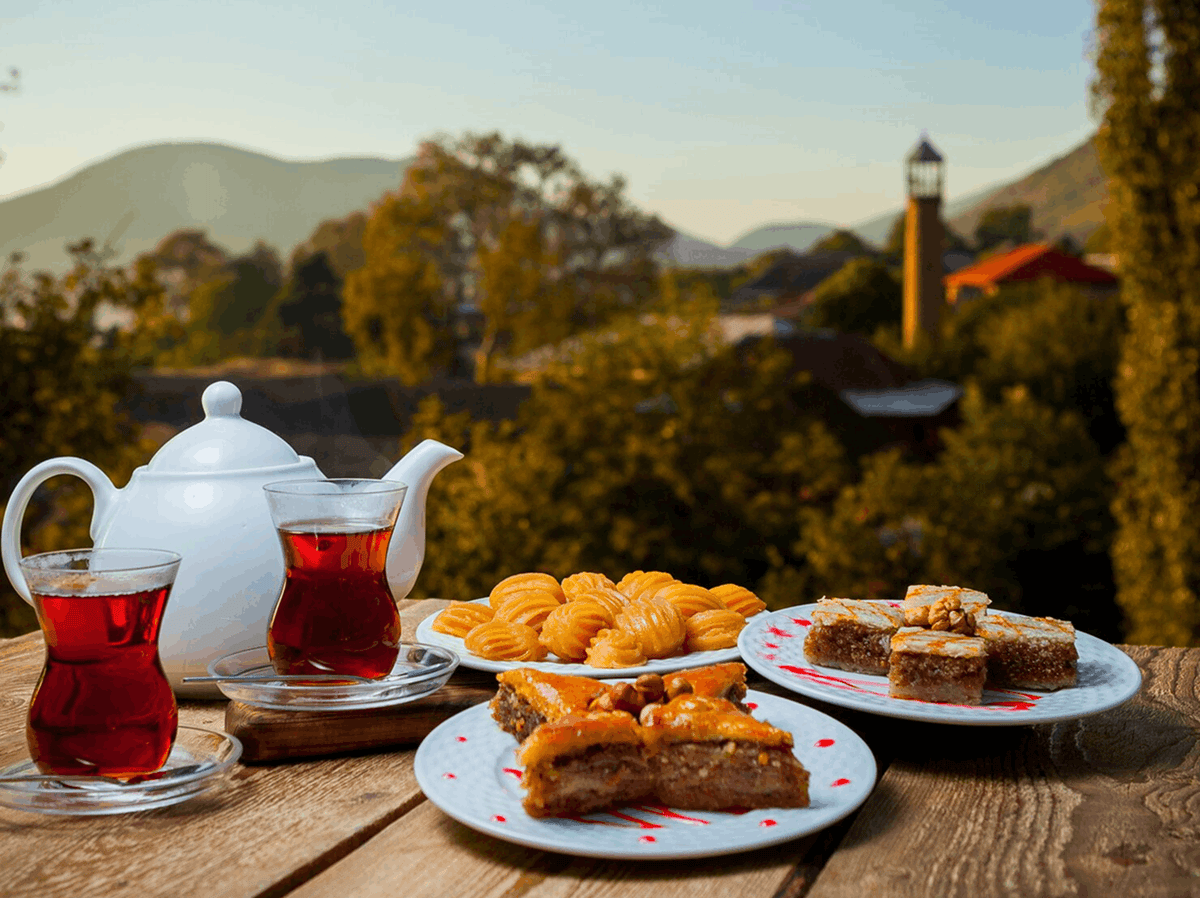 A white ceramic teapot and two glass cups filled with red tea are arranged on a wooden table alongside plates of pastries, set against a blurred outdoor landscape with hills and trees.