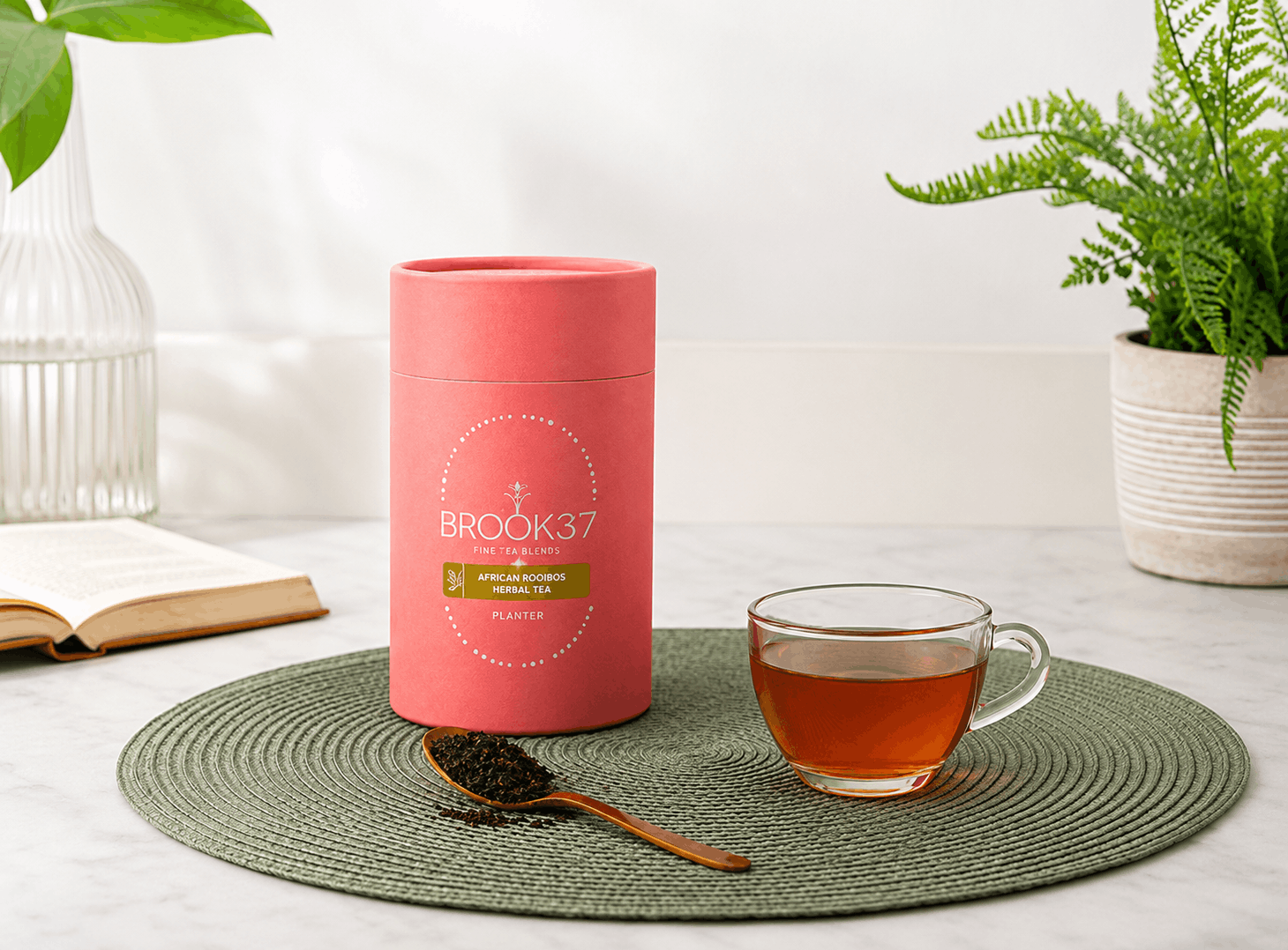 Brook37 Applause Orange herbal tea set with a pink cylindrical tin, a glass cup of amber tea, and a wooden spoon with loose tea leaves on a green woven placemat, styled on a white marble surface with a book and plants in the background.