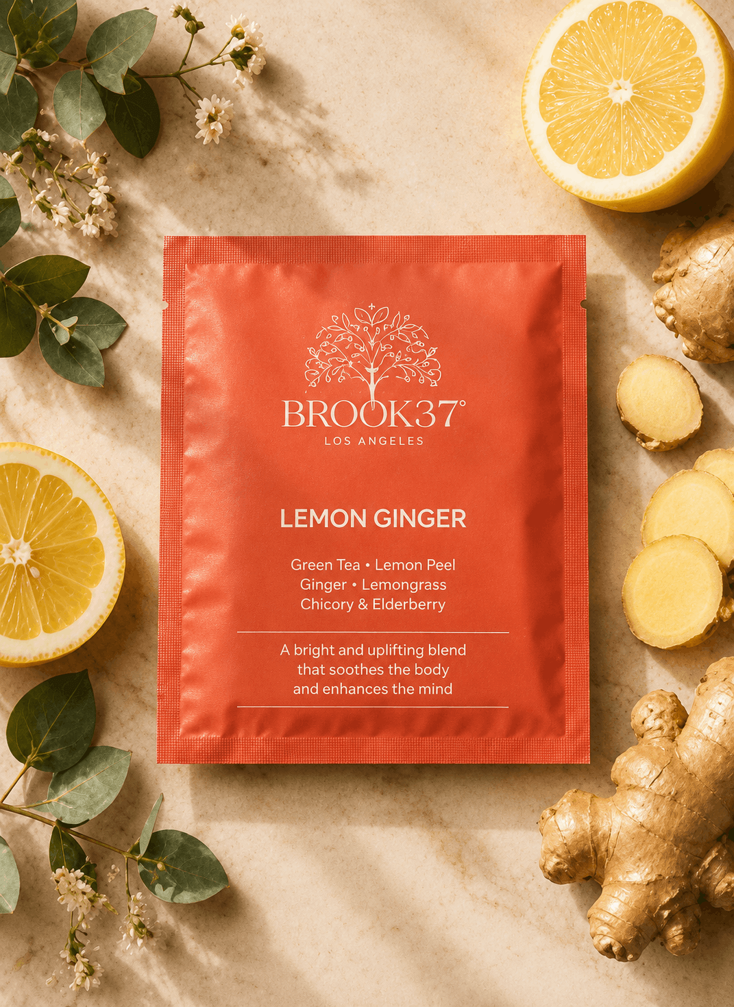 Brook37 Los Angeles Lemon Ginger green tea packet in a vibrant orange-red color, styled in a flat lay with fresh lemon slices, ginger root, and green leaves on a beige textured background.