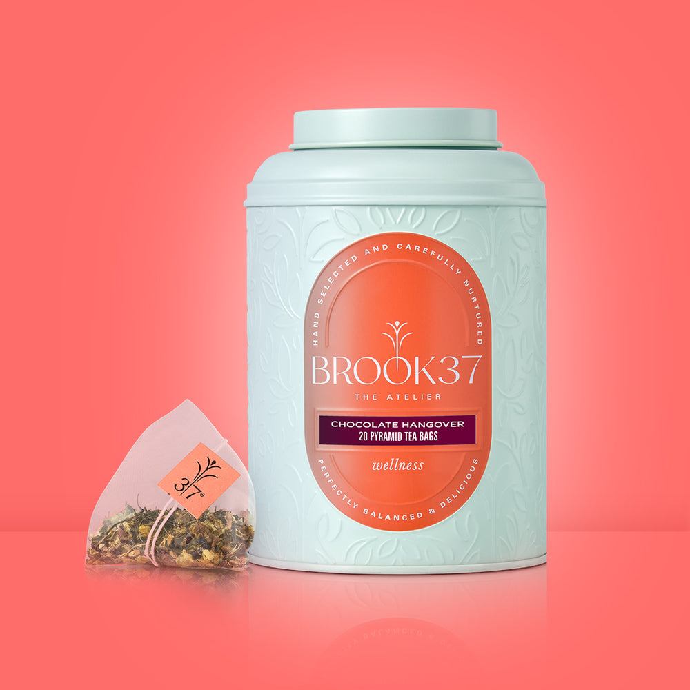 Product image prompt: The Chocolate Hangover tea in premium packaging beside a latte-style mug with frothy tea, cacao shells, cinnamon stick, ginger, and a few peppercorns styled minimally; warm, aromatic, mid-afternoon treat mood; diffused flattering light, film-inspired low contrast, ultra-realistic editorial product photography.